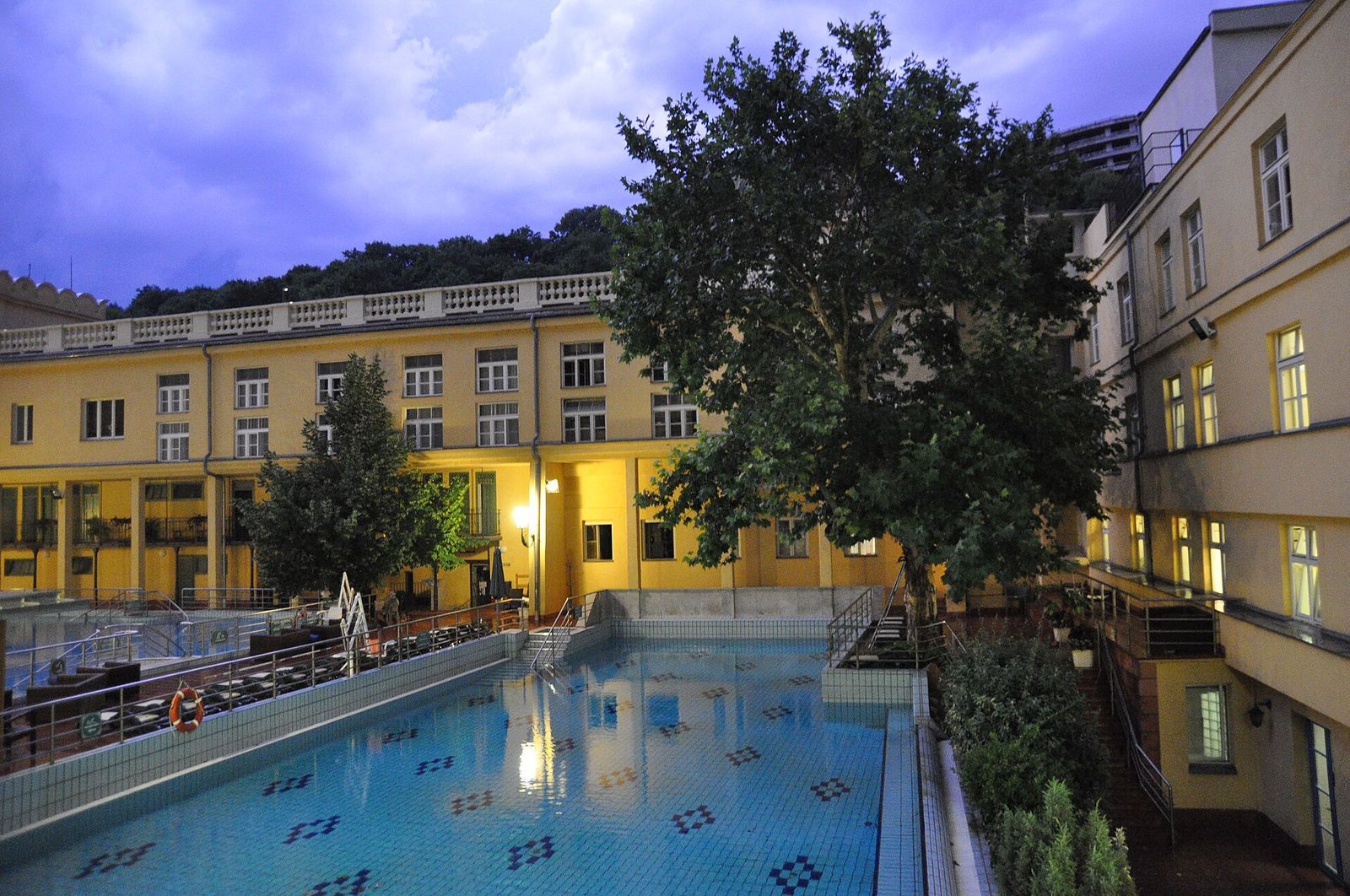 The outdoor swimming pool in the courtyard of Lukács Bath, framed by the bath's yellow buildings and plane trees